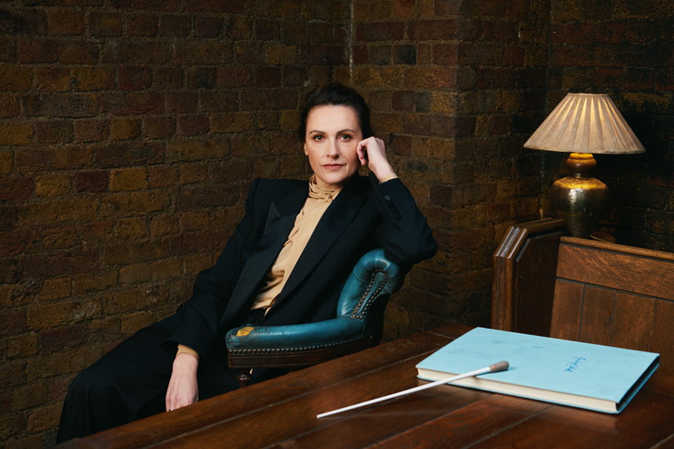 A woman with dark shoulder length hair, wearing a dark suit, leaning on a chair in front of a wooden desk.